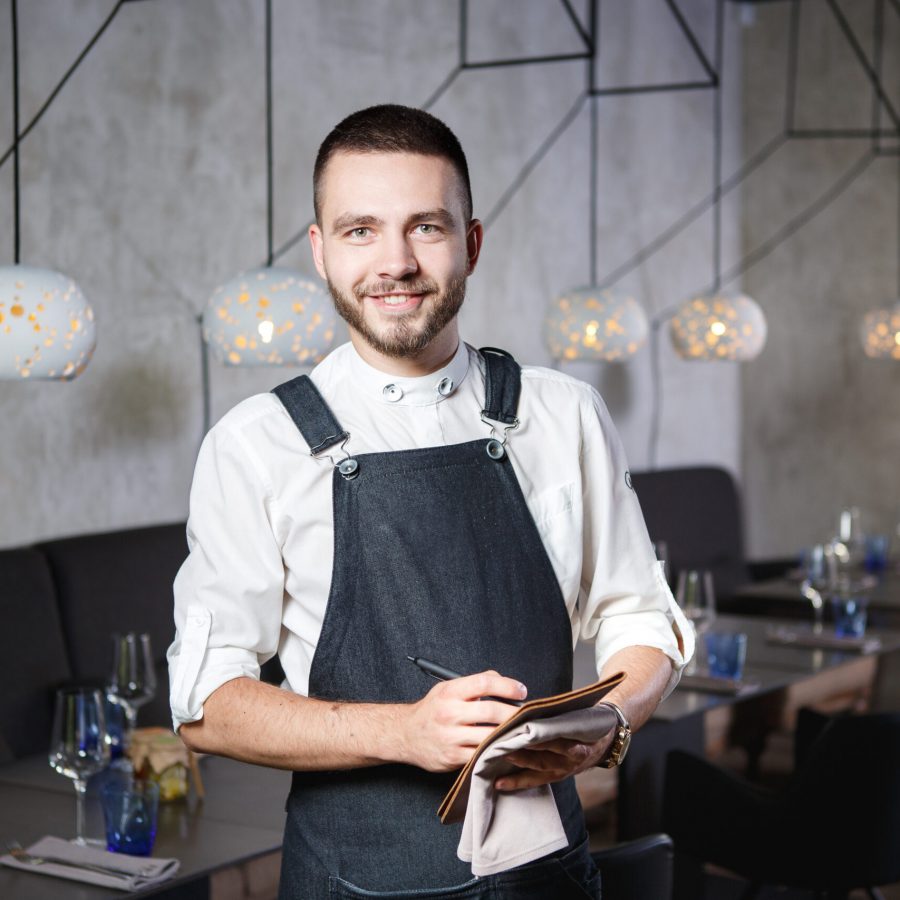 A young, smiling waiter in a restaurant, standing next to the tables with a glass of wine. Dressed in an apron, will take an order holding a notebook and a pen